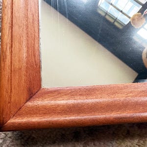 May include: Close-up of a rectangular mirror with a warm, reddish-brown wooden frame. The frame has a detailed, grooved design. The mirror reflects a partial view of a room with a window and a light-colored object.