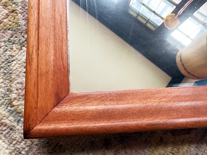 May include: Close-up of a rectangular mirror with a warm, reddish-brown wooden frame. The frame has a detailed, grooved design. The mirror reflects a partial view of a room with a window and a light-colored object.
