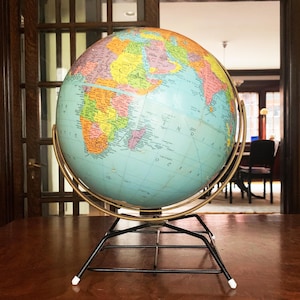 May include: A vintage globe with a colorful map of the world, mounted on a black metal stand with a gold-colored meridian ring. The globe is set on a dark wooden surface, with a blurred interior visible in the background.