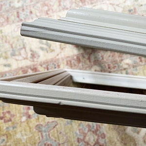 May include: Close-up of the corner of a white picture frame with a simple, ornate design. The frame is made of wood and has a smooth, painted finish.