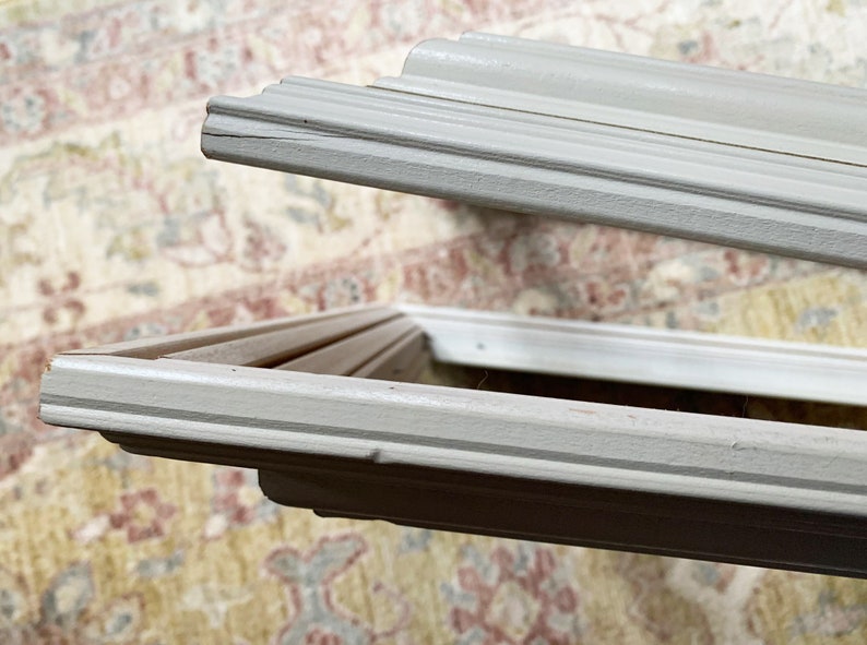May include: Close-up of the corner of a white picture frame with a simple, ornate design. The frame is made of wood and has a smooth, painted finish.