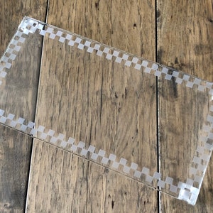 May include: A clear acrylic tray with a checkered pattern around the edge. The tray is on a wooden surface.