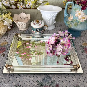 May include: A rectangular mirrored tray with gold-toned corner accents. The tray reflects a landscape painting with pink flowers and trees. Various decorative items, including a white vase and a small teacup, are arranged around the tray.