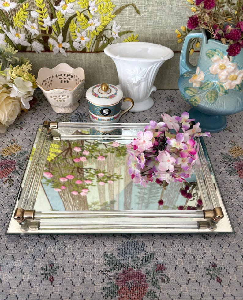 May include: A rectangular mirrored tray with gold-toned corner accents. The tray reflects a landscape painting with pink flowers and trees. Various decorative items, including a white vase and a small teacup, are arranged around the tray.