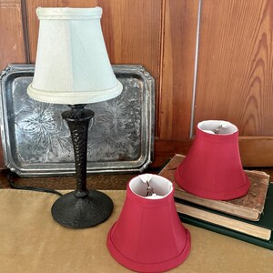 Op de afbeelding: Een kleine tafellamp met een lichtblauwe lampenkap en een zwarte voet. Twee rode lampenkappen staan naast de lamp. De lamp en kappen staan op een tafel met een zilveren dienblad en boeken op de achtergrond.