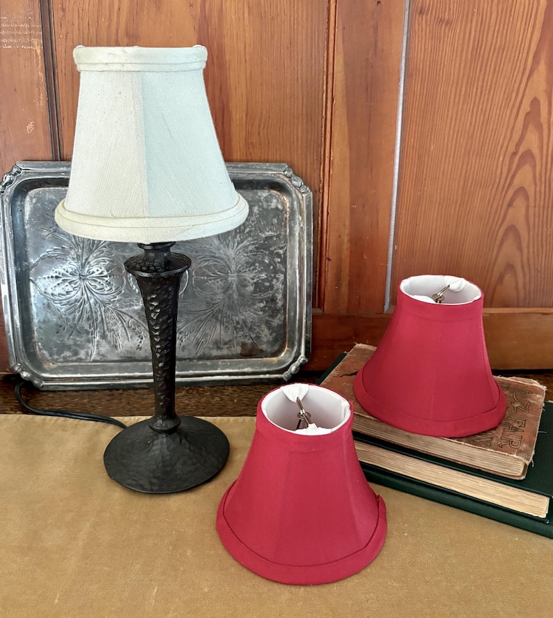 Op de afbeelding: Een kleine tafellamp met een lichtblauwe lampenkap en een zwarte voet. Twee rode lampenkappen staan naast de lamp. De lamp en kappen staan op een tafel met een zilveren dienblad en boeken op de achtergrond.