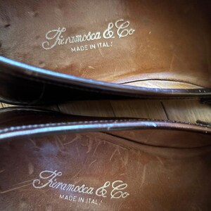 May include: A pair of dark brown leather loafers with a smooth finish. The interior insoles are tan leather and feature the white printed text "Fieramosca & Co" followed by "MADE IN ITALY" centered on the heel area of each shoe.