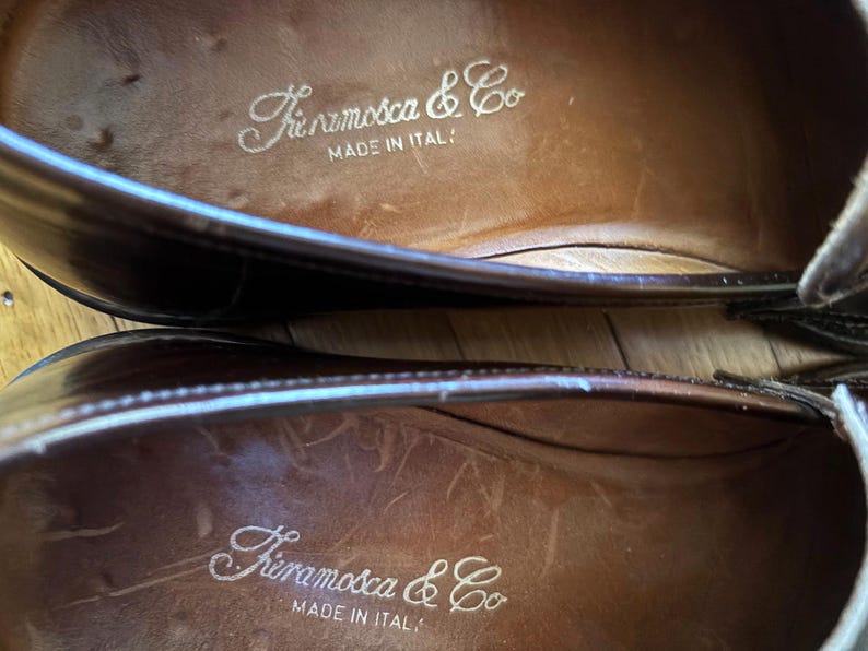 May include: A pair of dark brown leather loafers with a smooth finish. The interior insoles are tan leather and feature the white printed text "Fieramosca & Co" followed by "MADE IN ITALY" centered on the heel area of each shoe.
