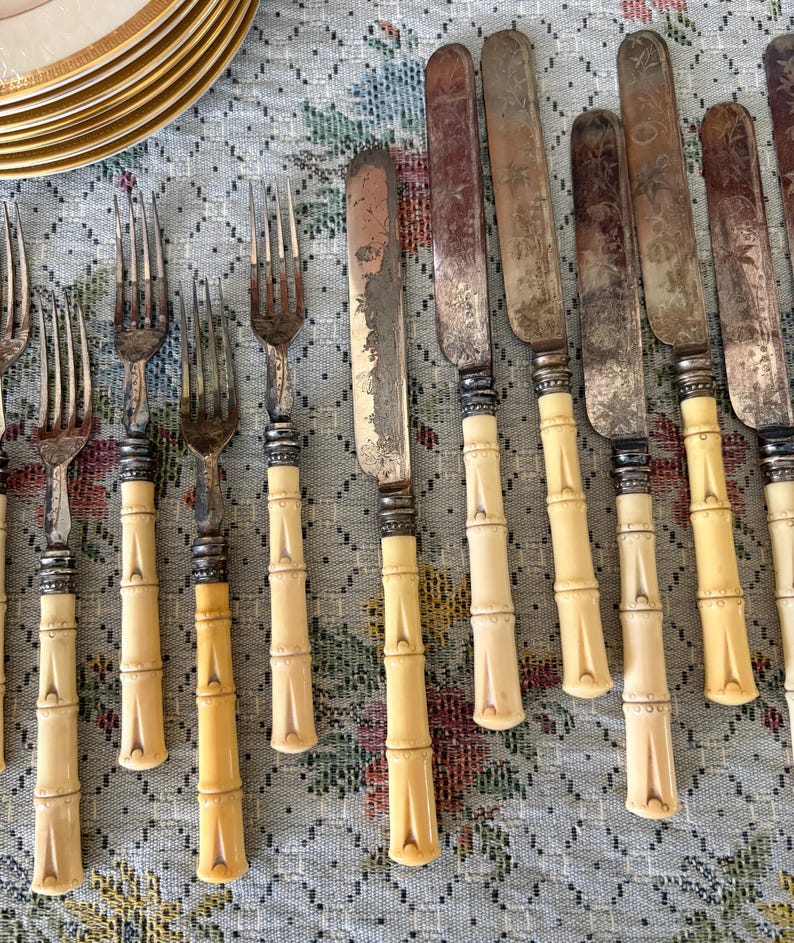 Pode incluir: Conjunto de talheres antigos com garfos e facas com cabos em estilo bambu. As l&acirc;minas das facas mostram sinais de desgaste. Os talheres est&atilde;o dispostos sobre uma toalha de mesa com padr&atilde;o floral.