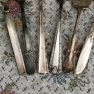 K&ouml;nnte beinhalten: Ein Set aus sieben Vintage-Silber-Gabeln mit verzierten Designs. Die Gabeln sind in einer Reihe auf einer gemusterten Stoffoberfl&auml;che angeordnet.