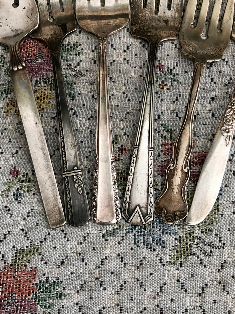 K&ouml;nnte beinhalten: Ein Set aus sieben Vintage-Silber-Gabeln mit verzierten Designs. Die Gabeln sind in einer Reihe auf einer gemusterten Stoffoberfl&auml;che angeordnet.