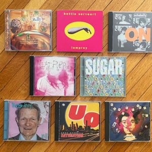 May include: A collection of nine compact discs (CDs) with various album covers. The CDs are arranged in three rows, with three CDs in each row. The CDs are all different colors and have different designs. The CDs are all in plastic cases.