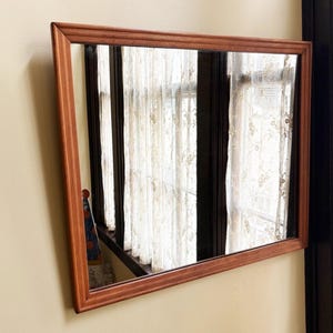 May include: A rectangular mirror with a brown wooden frame is mounted on a beige wall. The mirror reflects a window with white curtains and a glimpse of a patterned object. The frame has a simple, classic design.
