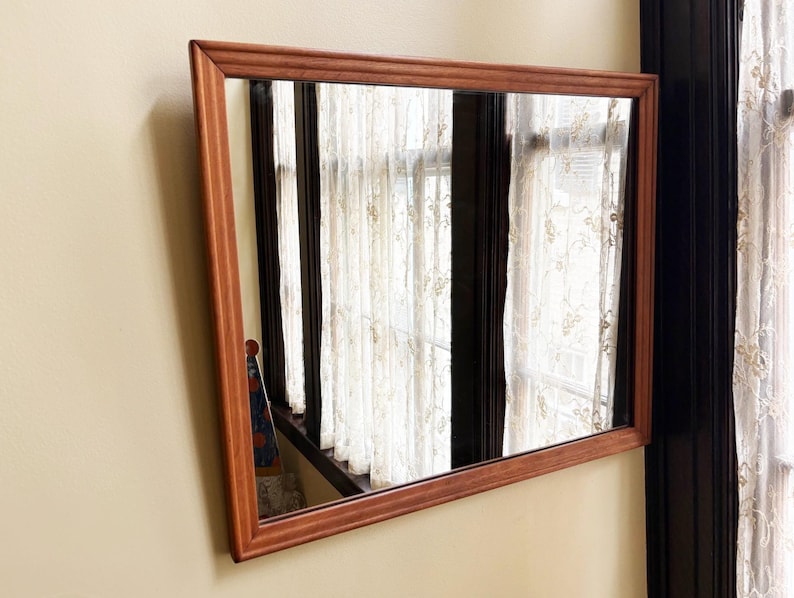 May include: A rectangular mirror with a brown wooden frame is mounted on a beige wall. The mirror reflects a window with white curtains and a glimpse of a patterned object. The frame has a simple, classic design.