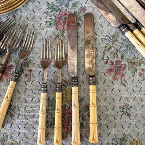 Pode incluir: Conjunto de talheres antigos com garfos e facas. As facas t&ecirc;m l&acirc;minas prateadas e os garfos t&ecirc;m dentes prateados. As pegas s&atilde;o de cor creme claro e em estilo bambu. Sobre uma toalha de mesa com padr&atilde;o floral.