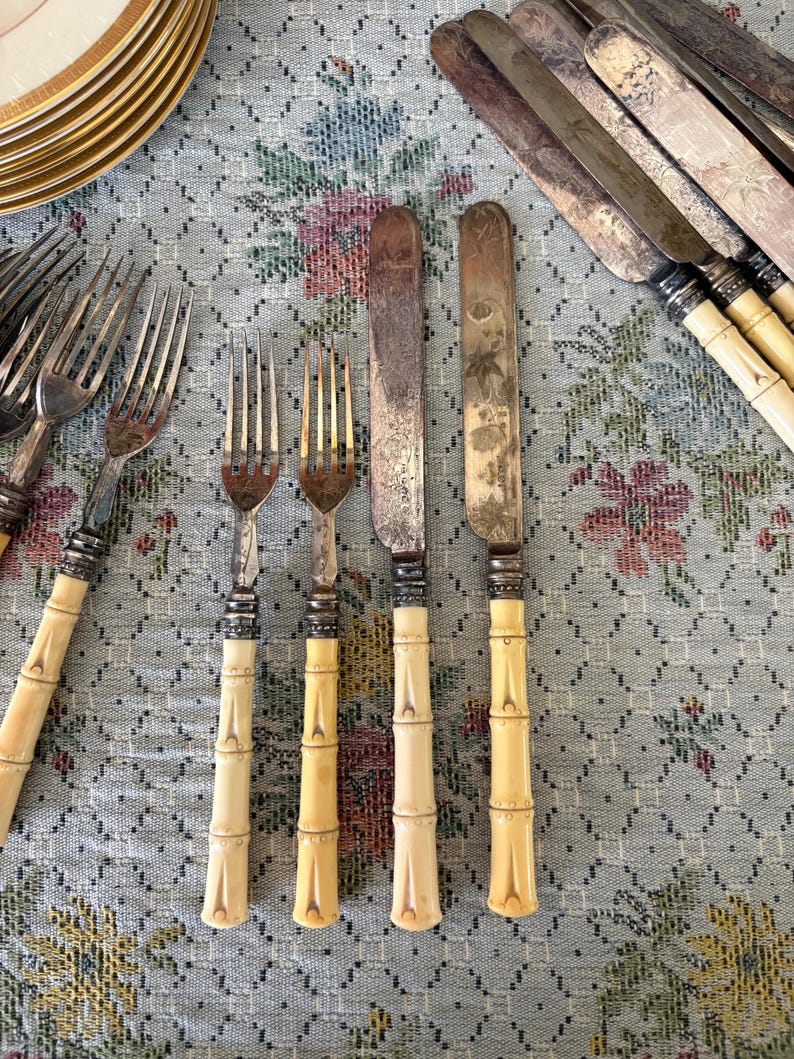 Pode incluir: Conjunto de talheres antigos com garfos e facas. As facas t&ecirc;m l&acirc;minas prateadas e os garfos t&ecirc;m dentes prateados. As pegas s&atilde;o de cor creme claro e em estilo bambu. Sobre uma toalha de mesa com padr&atilde;o floral.