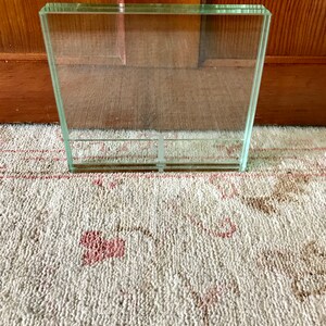 May include: A clear glass picture frame with a square shape. The frame is sitting on a white rug with a pink floral pattern.