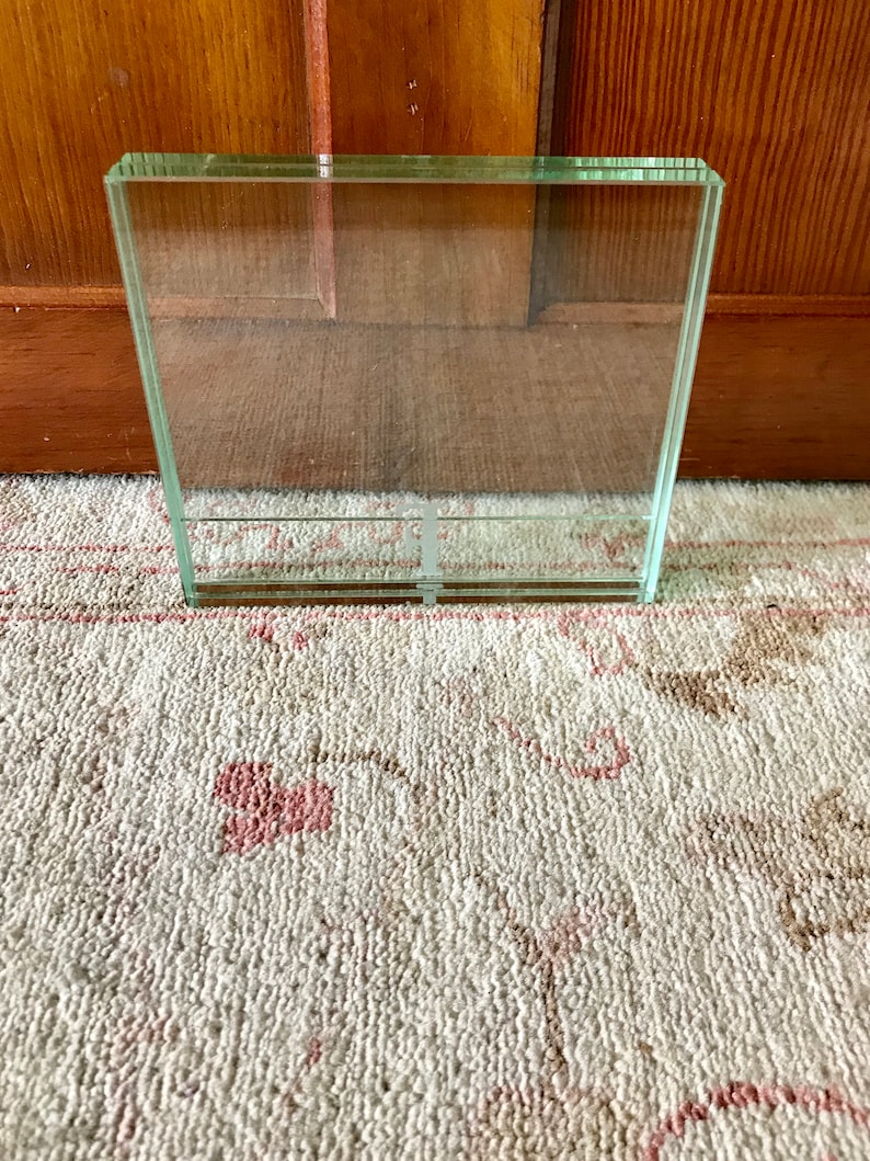 May include: A clear glass picture frame with a square shape. The frame is sitting on a white rug with a pink floral pattern.