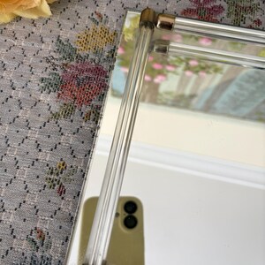 May include: A rectangular mirror with clear acrylic and gold-toned metal accents. The mirror's frame features clear acrylic rods and gold-toned metal corner pieces. The mirror reflects a blurred image of a room with pink flowers.