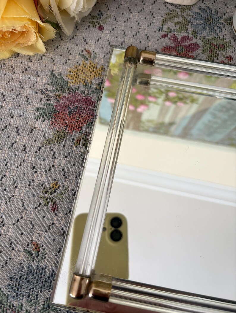 May include: A rectangular mirror with clear acrylic and gold-toned metal accents. The mirror's frame features clear acrylic rods and gold-toned metal corner pieces. The mirror reflects a blurred image of a room with pink flowers.