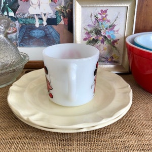 May include: A white ceramic mug with a black and red illustration of two girls wearing dresses and hats. The mug is sitting on a white saucer with a scalloped edge.