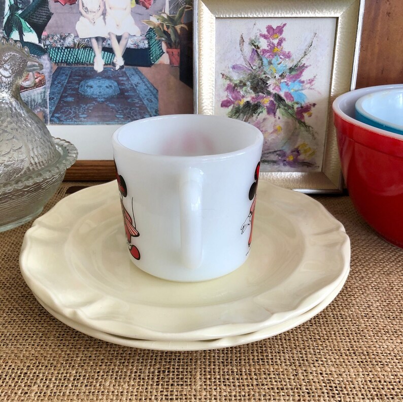 May include: A white ceramic mug with a black and red illustration of two girls wearing dresses and hats. The mug is sitting on a white saucer with a scalloped edge.
