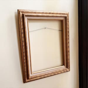 May include: A rectangular picture frame with an ornate, textured gold finish and a light beige inner border. The frame features a stepped profile and includes a thin metal hanging wire stretched across the back interior for wall mounting purposes.