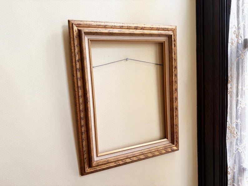 May include: A rectangular picture frame with an ornate, textured gold finish and a light beige inner border. The frame features a stepped profile and includes a thin metal hanging wire stretched across the back interior for wall mounting purposes.