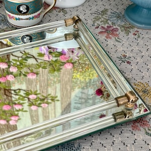 May include: Rectangular mirrored tray with clear glass rods and gold-toned corner accents. The tray reflects a landscape scene with pink flowers and trees. A decorative teacup and other vintage items are in the background.