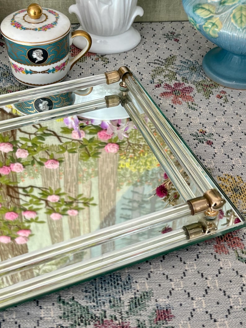 May include: Rectangular mirrored tray with clear glass rods and gold-toned corner accents. The tray reflects a landscape scene with pink flowers and trees. A decorative teacup and other vintage items are in the background.