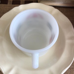 May include: A white ceramic mug with a scalloped rim sits on a white ceramic saucer. The mug has a white handle and a smooth, glossy finish.