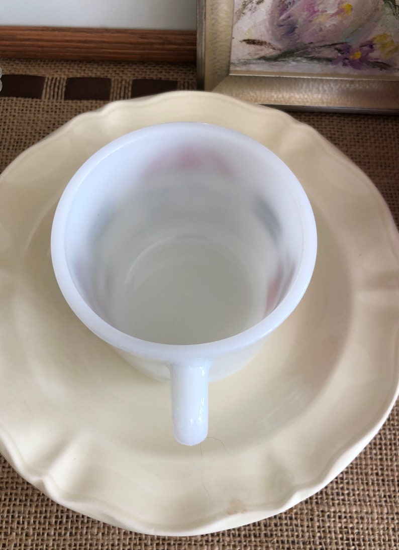 May include: A white ceramic mug with a scalloped rim sits on a white ceramic saucer. The mug has a white handle and a smooth, glossy finish.