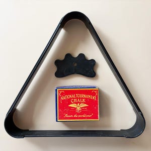 K&ouml;nnte beinhalten: Ein schwarzes Billard-Kugel-Dreieck, eine schwarze Br&uuml;cke und eine Schachtel "National Tournament Chalk" mit rotem und blauem Design. Die Kreideschachtel hat ein Adleremblem und den Text "Known the world over".