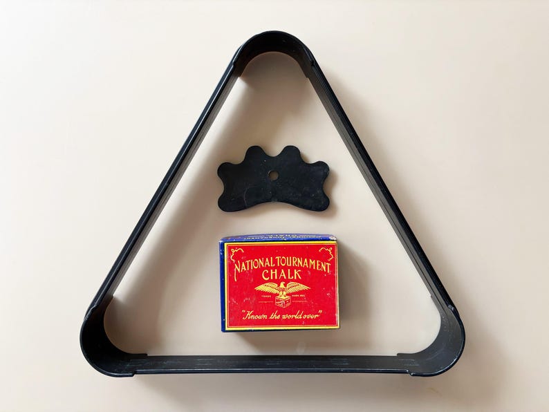 K&ouml;nnte beinhalten: Ein schwarzes Billard-Kugel-Dreieck, eine schwarze Br&uuml;cke und eine Schachtel "National Tournament Chalk" mit rotem und blauem Design. Die Kreideschachtel hat ein Adleremblem und den Text "Known the world over".