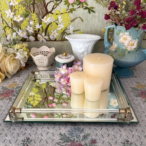 May include: A mirrored glass tray with gold-toned metal accents holds three ivory candles and a small bouquet of purple flowers. A variety of decorative vases and a floral tapestry are in the background, creating a vintage aesthetic.