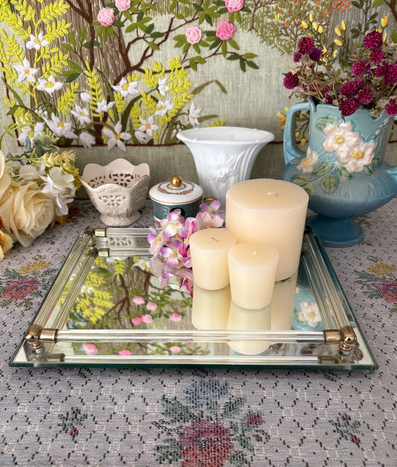 May include: A mirrored glass tray with gold-toned metal accents holds three ivory candles and a small bouquet of purple flowers. A variety of decorative vases and a floral tapestry are in the background, creating a vintage aesthetic.