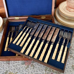 Pode incluir: Um conjunto de talheres vintage numa caixa de madeira com forro de veludo azul. O conjunto inclui garfos e facas com cabos creme em estilo bambu e l&acirc;minas prateadas ornamentadas. A caixa est&aacute; aberta, revelando a disposi&ccedil;&atilde;o dos utens&iacute;lios.