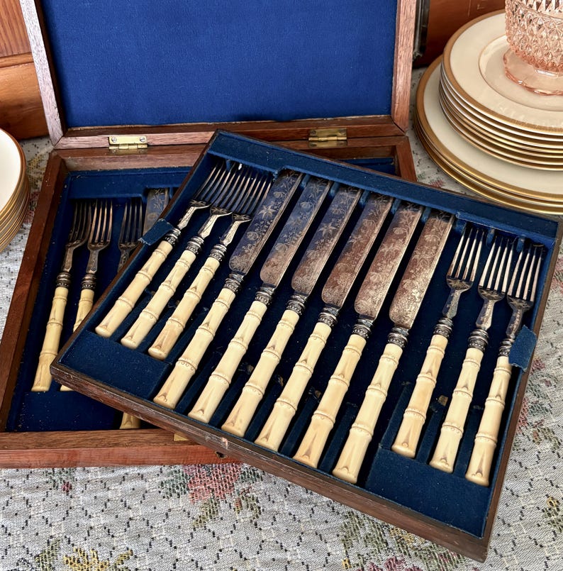 Pode incluir: Um conjunto de talheres vintage numa caixa de madeira com forro de veludo azul. O conjunto inclui garfos e facas com cabos creme em estilo bambu e l&acirc;minas prateadas ornamentadas. A caixa est&aacute; aberta, revelando a disposi&ccedil;&atilde;o dos utens&iacute;lios.