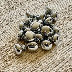 May include: A pile of chrome cabinet knobs, with a shiny, reflective finish. The knobs are round and have a simple design.