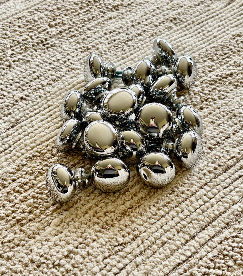 May include: A pile of chrome cabinet knobs, with a shiny, reflective finish. The knobs are round and have a simple design.