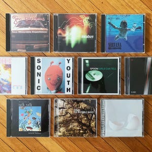 May include: A collection of 10 compact discs (CDs) with various album covers. The covers feature a variety of colors, designs, and band names, including "Gin Blossoms", "Incubus", "Nirvana", "Sonic Youth", "Spoon", "Talk Talk", and "The Invisible Band".