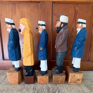May include: Five carved wooden sailor figurines. Each figure is standing on a small wooden block. The sailors are wearing coats, hats, and some have pipes. The coats are blue, yellow, and brown. The hats are white.