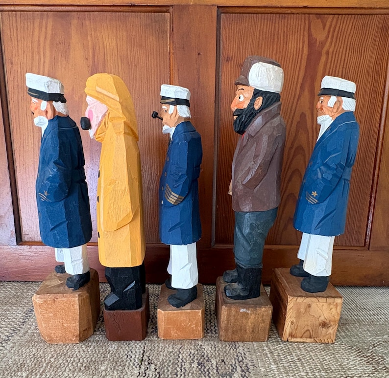 May include: Five carved wooden sailor figurines. Each figure is standing on a small wooden block. The sailors are wearing coats, hats, and some have pipes. The coats are blue, yellow, and brown. The hats are white.