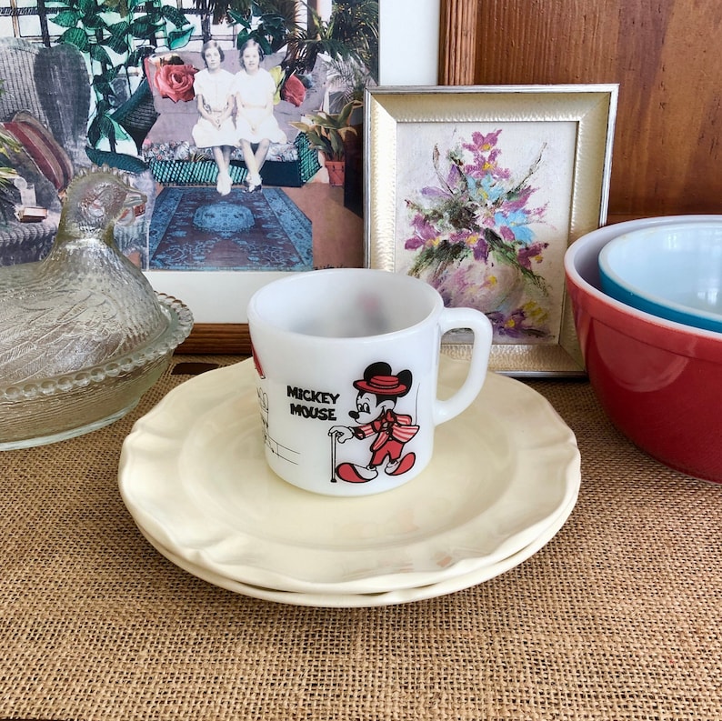 May include: A white ceramic mug with a Mickey Mouse illustration. The mug has a red handle and says 'Mickey Mouse' on the front. The mug is sitting on a white saucer with a scalloped edge.