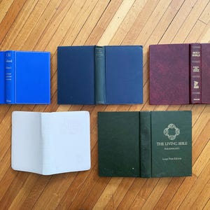 Op de afbeelding: Een assortiment hardcover boeken in verschillende kleuren, waaronder blauw, marineblauw, bordeauxrood en groen, wordt open en dicht getoond op een houten oppervlak. Sommige boeken hebben tekst op de rug en de kaft, zoals "Holy Bible" en "The Living Bible."