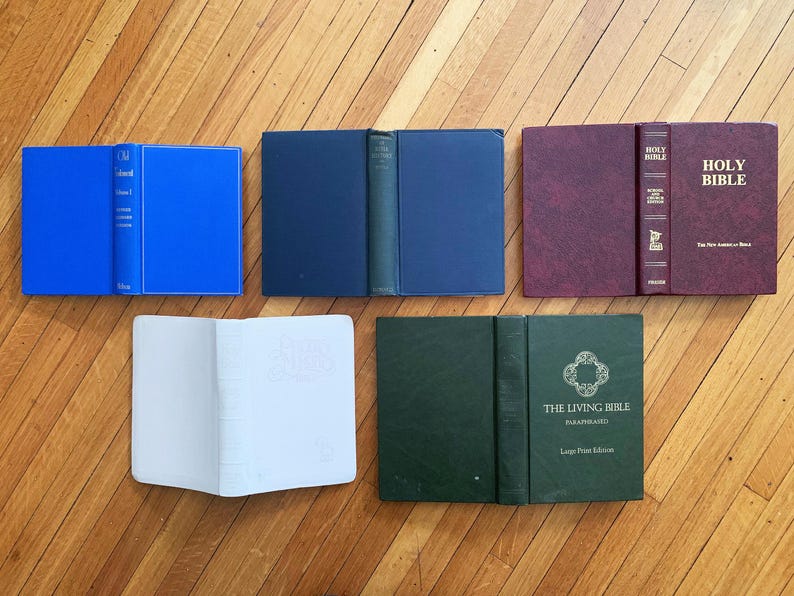 Op de afbeelding: Een assortiment hardcover boeken in verschillende kleuren, waaronder blauw, marineblauw, bordeauxrood en groen, wordt open en dicht getoond op een houten oppervlak. Sommige boeken hebben tekst op de rug en de kaft, zoals "Holy Bible" en "The Living Bible."