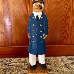 May include: A hand-carved wooden figurine of a sea captain. The captain wears a navy blue coat with gold buttons and stripes, a white hat with a gold anchor, and white pants. The figurine stands on a small wooden base.