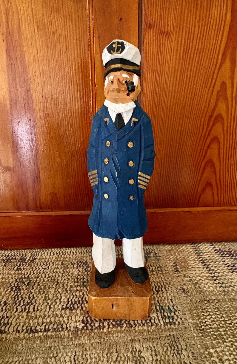 May include: A hand-carved wooden figurine of a sea captain. The captain wears a navy blue coat with gold buttons and stripes, a white hat with a gold anchor, and white pants. The figurine stands on a small wooden base.
