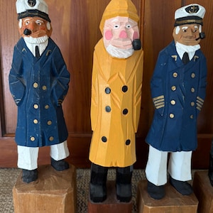 May include: Three hand-carved wooden sailor figurines. The figures wear blue coats, white pants, and sailor hats. One figure wears a yellow raincoat. Each figure has a pipe in their mouth and stands on a wooden block base.