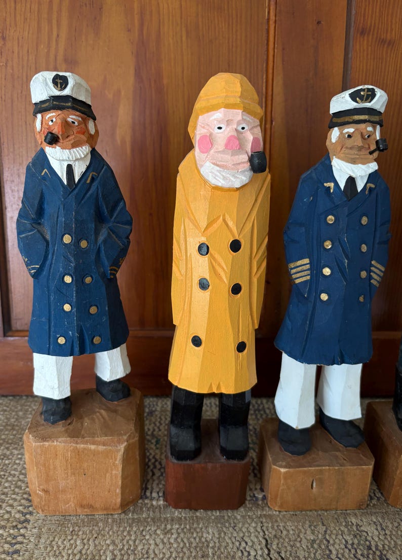 May include: Three hand-carved wooden sailor figurines. The figures wear blue coats, white pants, and sailor hats. One figure wears a yellow raincoat. Each figure has a pipe in their mouth and stands on a wooden block base.